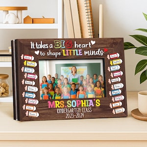 Op de afbeelding: Een houten fotolijst met de tekst "It takes a BIG heart to shape LITTLE minds." De lijst bevat een foto van een leraar en leerlingen. De lijst bevat ook de tekst "MRS. SOPHIA'S KINDERGARTEN CLASS 2023-2024."