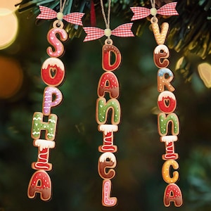May include: Three gingerbread-style Christmas ornaments spelling out the names "Sophia", "Daniel", and "Veronica". Each letter is decorated with icing and attached to a string with a red and white checkered bow.