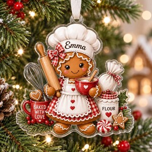 May include: A clear acrylic Christmas ornament featuring a gingerbread person wearing a chef hat with the name Emma, a red and white gingham apron, holding a rolling pin and a red bowl, with a red mug labeled Baked With Love and a flour jar.