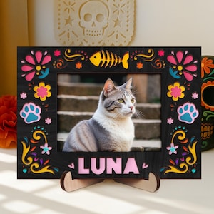 May include: A black photo frame with colorful floral and paw print designs, and the name "LUNA" in pink letters. The frame holds a photo of a gray and white cat. A wooden stand supports the frame.