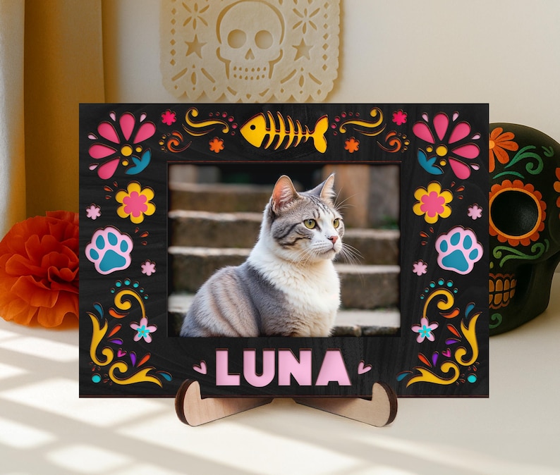 May include: A black photo frame with colorful floral and paw print designs, and the name "LUNA" in pink letters. The frame holds a photo of a gray and white cat. A wooden stand supports the frame.