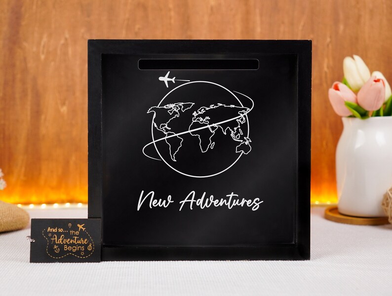 May include: Black wooden box with a slot on the top. The box has a white outline of the world with an airplane flying above it. The text "New Adventures" is written below the world.