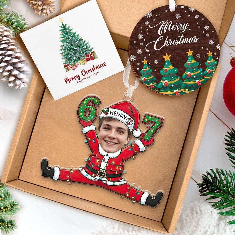 May include: A Christmas-themed gift box containing a personalized ornament with a photo, a wooden ornament with "Merry Christmas" text, and a card with a Christmas tree design. The personalized ornament features a person's face in a Santa suit.