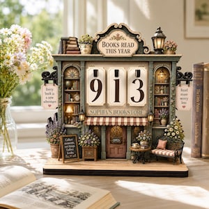May include: A decorative tabletop bookstore display featuring a flip-style counter displaying the number 913. The structure includes miniature bookshelves, a central door with an open sign, floral accents, and signs reading Books Read This Year, Sophia's Bookstore, Every book is a new adventure, Read more worry less, and Collect moments, not things. Collect books.
