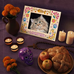 May include: A decorative wooden picture frame with a cat photo, the name "Lucifer", and colorful floral and paw print accents. The scene includes candles, a purple skull, apples, and bread, suggesting a Day of the Dead theme.