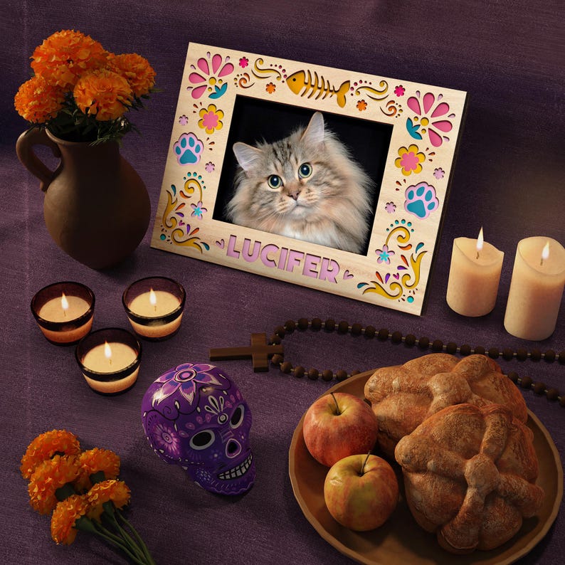 May include: A decorative wooden picture frame with a cat photo, the name "Lucifer", and colorful floral and paw print accents. The scene includes candles, a purple skull, apples, and bread, suggesting a Day of the Dead theme.