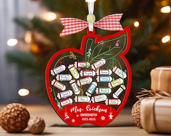 May include: A red apple-shaped ornament featuring a clear center filled with small pencil-shaped name tags. The ornament includes a red and white gingham bow, a white ribbon hanger, and text reading Mrs. Erickson KINERGARTEN 2025-2026.