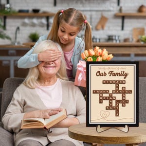 May include: A framed print with a beige background and black frame. The print features a crossword puzzle with the words "Our Family" at the top. The crossword puzzle is made up of names, including Richard, Frank, Daniel, Brian, Lucy, Katy, and Layla. The print has a brown heart at the bottom.