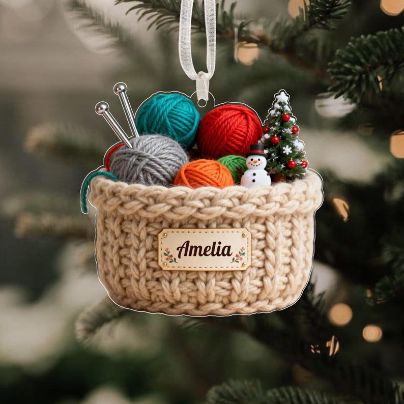 Puede incluir: Adorno navide&ntilde;o de cesta tejida beige llena de bolas de hilo de colores, agujas de tejer, un &aacute;rbol de Navidad en miniatura y un mu&ntilde;eco de nieve. La cesta tiene una peque&ntilde;a etiqueta rectangular que dice "Amelia".