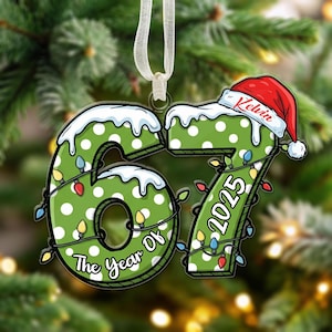 May include: A Christmas tree ornament with the numbers 67 and 2025 in green with white polka dots. The numbers are decorated with fairy lights and snow. A red Santa hat with the name "Kelvin" is on top. The ornament hangs from a white ribbon.