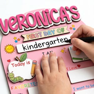 May include: Pink wooden sign with the text "VERONICA'S FIRST DAY OF kindergarten". The sign has decorative elements including a sun, stars, an apple, a pencil, and a caterpillar. A hand is writing on the sign with a black marker.