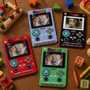 Op de afbeelding: Vier kleurrijke fotolijsten in de vorm van een gameconsole. Elke lijst bevat een foto van een vader en kind, met de woorden "Daddy" of "Papa" bovenaan. De lijsten zijn rood, blauw, groen en zwart, met gamecontrollerknoppen en de tekst "THANKS FOR ALWAYS LEVELING US UP!"