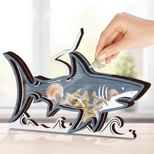 May include: A wooden shark-shaped ornament with a blue-grey finish, brown trim, and white accents. The shark's interior is filled with seashells and shark teeth. A hand is placing a shark tooth into the shark's mouth. The base features wave-like cutouts.