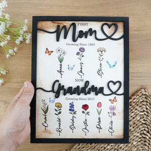 May include: Black framed wood sign with the words "FIRST Mom Growing Since 1993" and "NOW Grandma Growing Since 2017" with a heart symbol after each word. The sign has a vintage background with flowers and butterflies and names written below each flower.