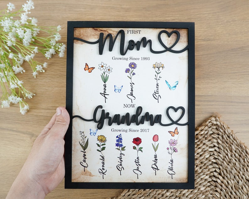 May include: Black framed wood sign with the words "FIRST Mom Growing Since 1993" and "NOW Grandma Growing Since 2017" with a heart symbol after each word. The sign has a vintage background with flowers and butterflies and names written below each flower.