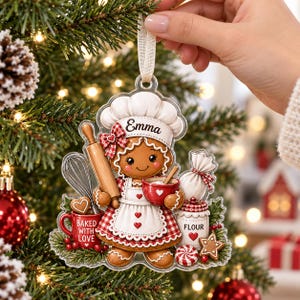 May include: A clear acrylic Christmas ornament shaped like a gingerbread person wearing a chef hat with the name Emma, holding a rolling pin and a red bowl. The design includes a whisk, a red mug labeled Baked With Love, and a flour jar.