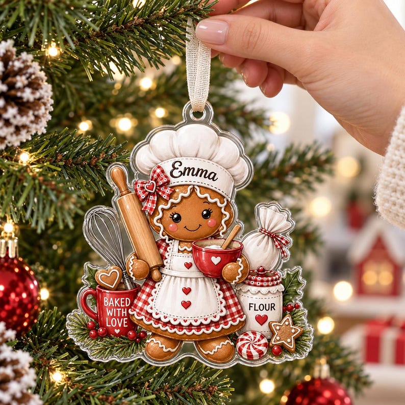 May include: A clear acrylic Christmas ornament shaped like a gingerbread person wearing a chef hat with the name Emma, holding a rolling pin and a red bowl. The design includes a whisk, a red mug labeled Baked With Love, and a flour jar.