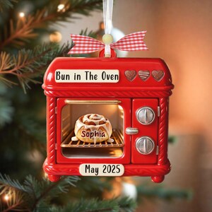Può includere: Ornamento natalizio a forma di forno rosso con la scritta "Bun in The Oven" e "May 2025". All'interno del forno c'è un rotolo di cannella con il nome "Sophia". L'ornamento ha un fiocco a scacchi rosso e bianco.