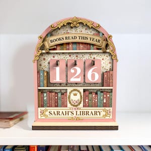 May include: A pink wooden bookshelf-shaped display with the text "BOOKS READ THIS YEAR" and "126". The display has shelves with miniature book decorations and the text "SARAH'S LIBRARY".
