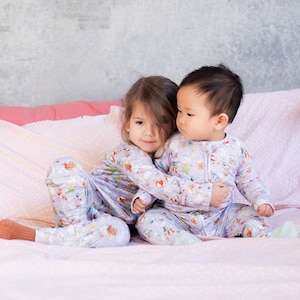 Op de afbeelding: Twee kinderen in bijpassende pyjama's met een bosdierprint zitten op een roze bed met witte stippen. De kinderen knuffelen elkaar en lachen.