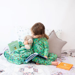 May include: A young child wearing green pajamas with a Peanuts character print is sitting on a bed with a stuffed animal and reading a book titled "The Adventures of Woodstock".