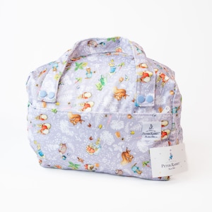 Pode incluir: Uma bolsa de mão de cor lilás claro com fundo branco e um padrão repetido de personagens do Peter Rabbit. A bolsa tem dois botões azuis de cada lado e um fecho de correr. A bolsa tem uma etiqueta que diz "The World of Peter Rabbit" e uma etiqueta menor que diz "Peter Rabbit".