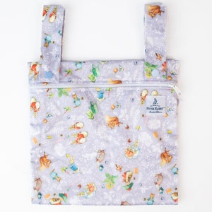 Pode incluir: Uma bolsa húmida reutilizável roxa com fecho de correr e duas correias ajustáveis. A bolsa apresenta um padrão repetitivo de personagens do Peter Rabbit e desenhos florais. A bolsa tem uma etiqueta branca com o texto "The World of Peter Rabbit" e um pequeno botão azul em cada alça.