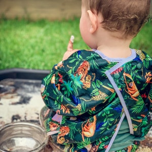 Könnte beinhalten: Ein Kleinkind trägt eine grüne, blaue und weiße gemusterte Lätzchen mit Tiger-, Affen- und Blumendesign. Das Lätzchen hat einen weißen Saum und einen Druckknopfverschluss.
