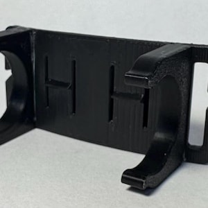 May include: A black plastic clip with a rectangular body and curved arms. The clip has two slots and two curved arms on each side. The clip is designed to hold objects. The clip is made of a matte black plastic.