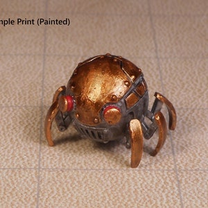 May include: A small, metallic, gold-painted spider-like robot with red eyes. The robot has a round body with a segmented design and six legs.