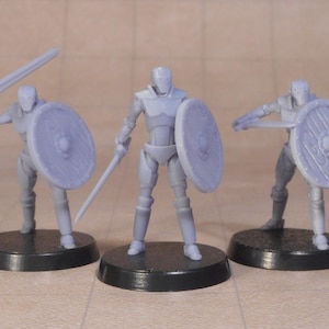 May include: Three gray miniature figures of warriors, each with a shield and sword. One figure holds a sword aloft, another at the ready, and the third is in a defensive stance. All are on black circular bases.