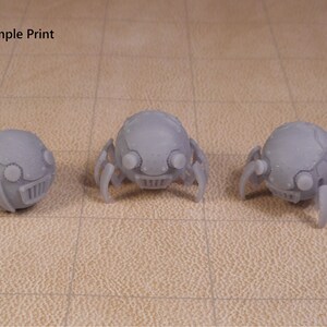 May include: Three 3D printed gray robot spiders with round bodies and six legs. The spiders have a detailed design with a face and eyes.
