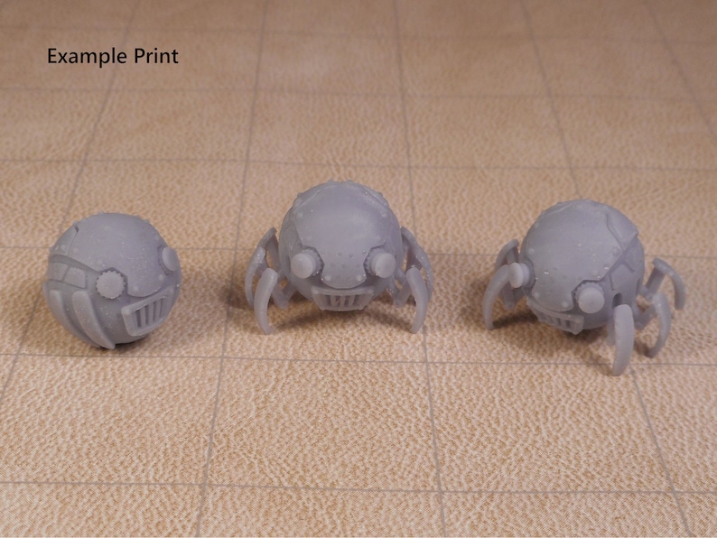 May include: Three 3D printed gray robot spiders with round bodies and six legs. The spiders have a detailed design with a face and eyes.