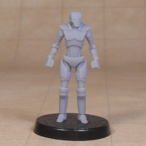 Pode incluir: Uma estatueta em miniatura cinza claro de um robô humanoide, em pé sobre uma base circular preta. O robô tem um design segmentado com placas semelhantes a armaduras no torso, braços e pernas. A figura tem cerca de 5 cm de altura.