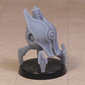 Puede incluir: Un modelo de robot en miniatura de color gris claro con una cabeza redondeada, rasgos circulares en forma de ojo y tres patas. El robot está montado sobre una base circular negra. El modelo parece ser de plástico.
