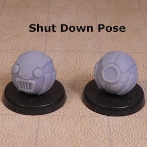 May include: A close-up of a small, round, gray robot toy in a shut-down pose. The robot has a face with two large eyes and a mouth with a grill-like design. The robot is sitting on a black base.