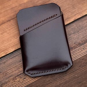 May include: A dark brown leather card holder with a stitched design. The rectangular wallet has a flap closure and is set against a wooden background. The leather has a glossy finish.
