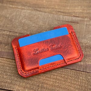 May include: A red leather cardholder with blue card slots. The cardholder has the words "Leather Tactics" embossed on the front. The edges are stitched with black thread. The cardholder is on a wooden surface.
