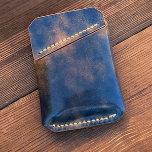 May include: A rectangular leather card holder featuring a marbled navy blue and brown finish with visible tan stitching along the edges and a diagonal pocket opening. The text "Each Marbled Shell Cordovan Pattern will be unique." is printed below.