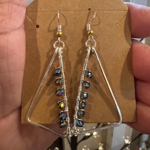 May include: A pair of silver-toned triangle earrings with iridescent beads. The earrings are suspended from silver hooks with gold-colored accents. The beads are wired to the bottom of the triangle frame.