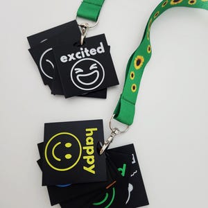 May include: A green lanyard with a sunflower pattern attached to two metal clips. Each clip holds a stack of square black plastic tags featuring various white or yellow emoji icons and text labels such as excited and happy in a sans-serif font.