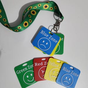 May include: A green lanyard with a sunflower pattern attached to a metal clip holding several square plastic tags. The tags are blue, green, red, and yellow, each featuring a white embossed face icon and text labels including Blue Zone, Green Zone, Red Zone, and Yellow Zone.