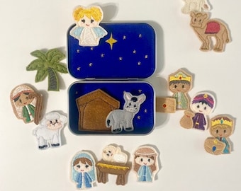 May include: A blue tin with a nativity scene inside, including a felt donkey, stable, and figures. Surrounding the tin are felt figures of an angel, palm tree, sheep, camel, and three wise men. The lid has a star design.