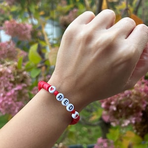 May include: Red beaded bracelet with white letter beads spelling out the word "RED" and red heart beads.