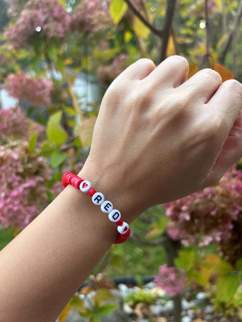 May include: Red beaded bracelet with white letter beads spelling out the word "RED" and red heart beads.