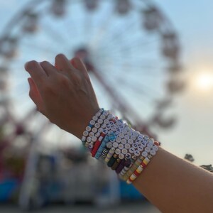 May include: A person's wrist with multiple colorful beaded bracelets. The bracelets have different colored beads and letters spelling out words like "RED", "HOPE", "MIDNIGHT", and "TTPOD".