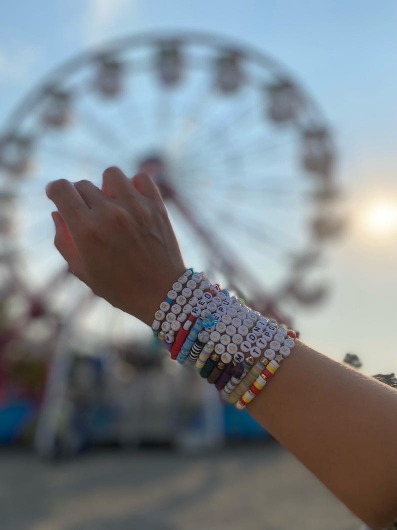 May include: A person's wrist with multiple colorful beaded bracelets. The bracelets have different colored beads and letters spelling out words like "RED", "HOPE", "MIDNIGHT", and "TTPOD".