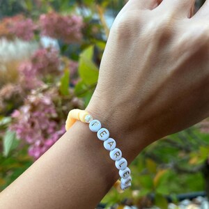May include: A white beaded bracelet with the word "FEARLESS" spelled out in gold letters. The bracelet is on a person's wrist and is made of two-toned beads, with a light yellow color on one side and a white color on the other.