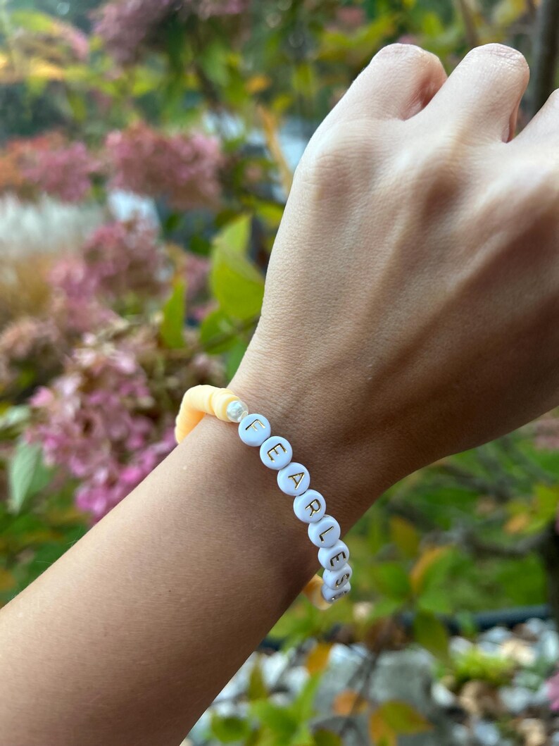 May include: A white beaded bracelet with the word "FEARLESS" spelled out in gold letters. The bracelet is on a person's wrist and is made of two-toned beads, with a light yellow color on one side and a white color on the other.