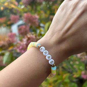 May include: A colorful beaded bracelet with the word "ERASE" spelled out in white letters on white beads. The bracelet is made with pastel blue, yellow, and pink beads.